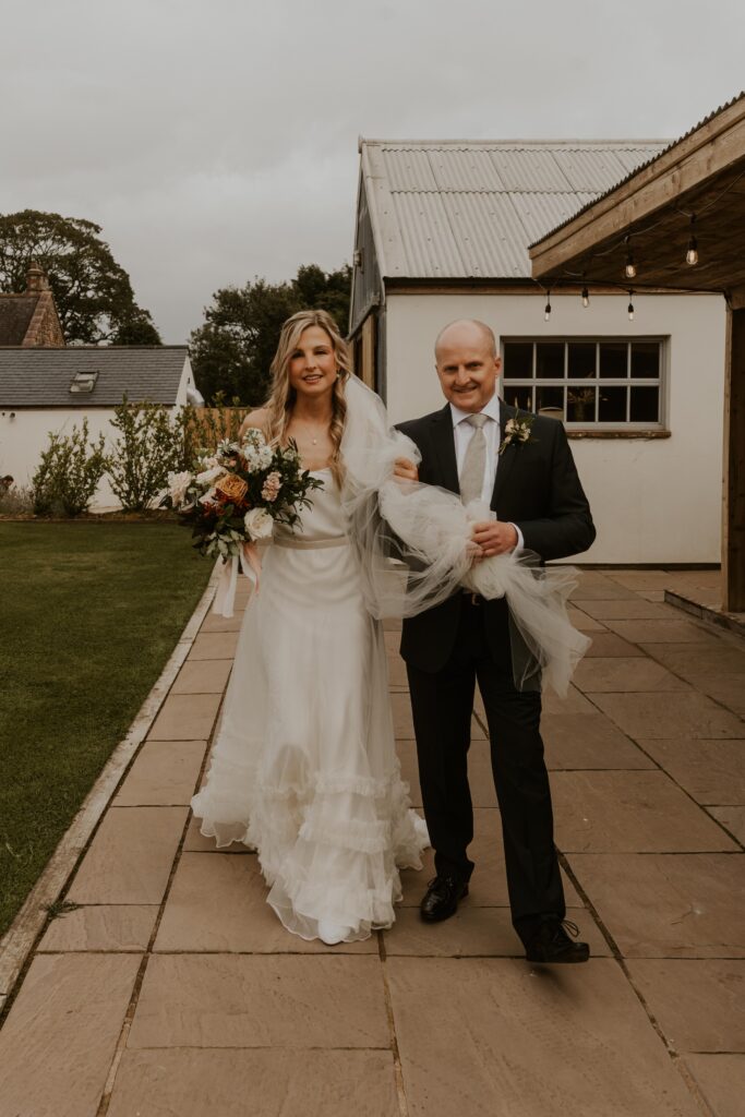 The bride and her Dad in beautiful half penny london wedding dress walking to the ceremony at Eden Barn wedding venue