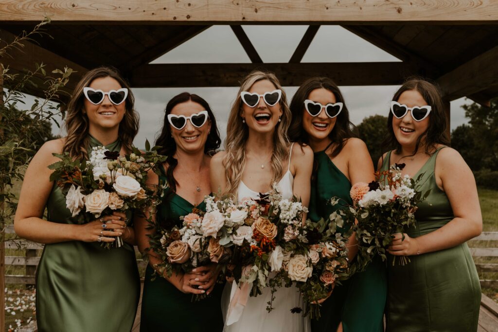 relaxed and fun wedding photographer capturing group shots of the bride enjoying her time with her bridesmaids in green silk dresses with heart sunglasses, smiling and having fun