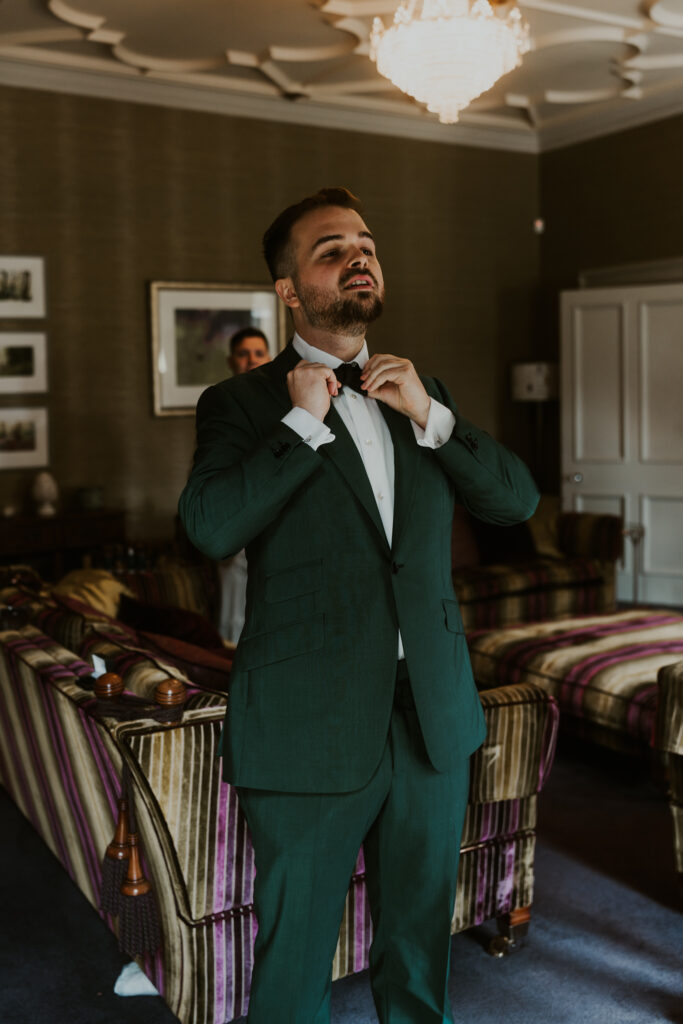 The groom tieing his bow tie in green suit ahead of his amazing wedding at tithe barn 